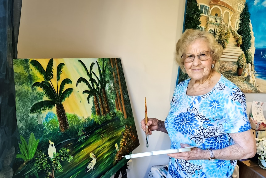 An older woman in a bright outfit paints a tropical scene on a canvas.