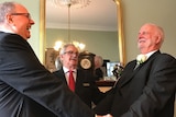 Father Rob Whalley and Father John Davis in black wedding suits, facing each other holding hands and smiling.