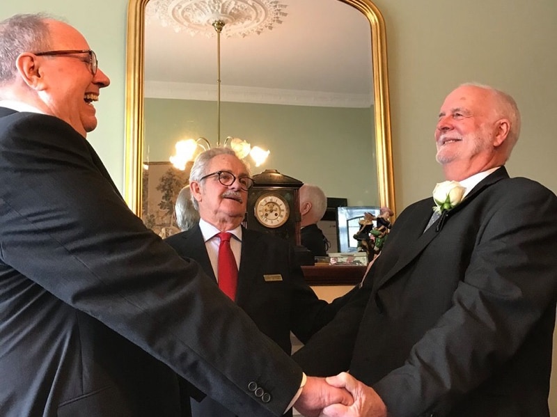 Father Rob Whalley and Father John Davis in black wedding suits, facing each other holding hands and smiling.