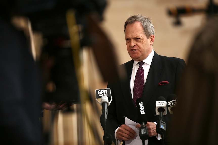 A man wearing a suit and tie frowns as he speaks before microphones. In the foreground is camera equipment.
