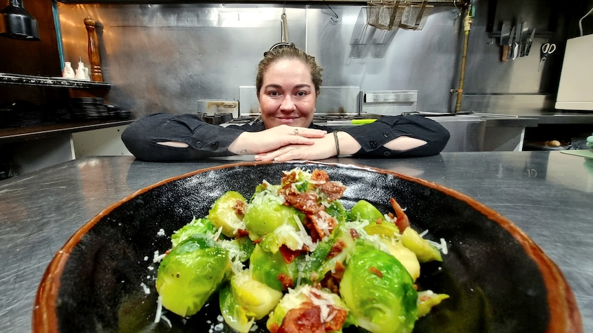 Young woman rests her chin on her hands and smiles at camera. In front of her is a plate of brussels sprouts