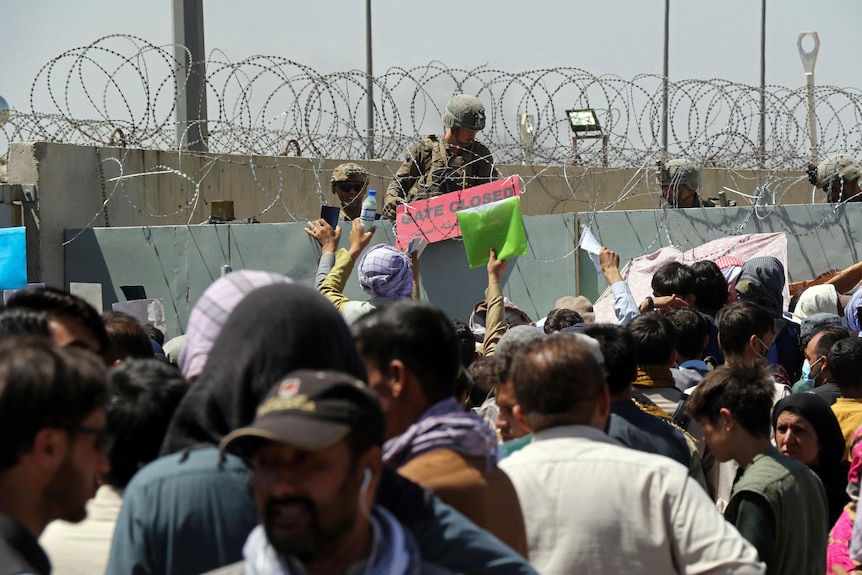 A US soldier holds a sign indicating a gate is closed as hundreds of people gather some holding documents.