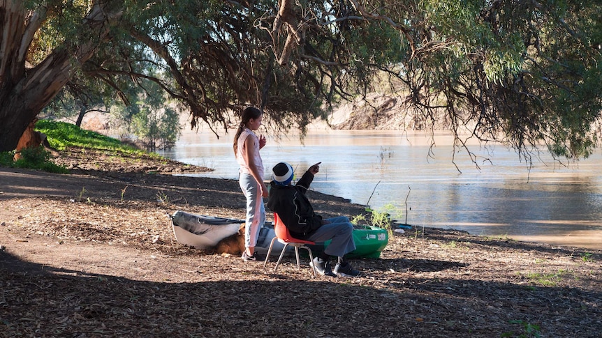 A man sits in a chair next to a river, pointing something out to a young girl standing next to a kayak.