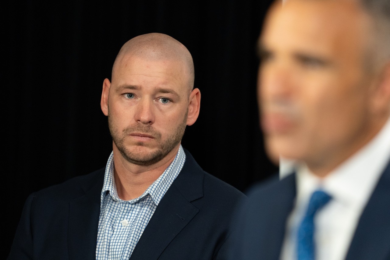 Un hombre en foco escucha al primer ministro Peter Malinauskas, que está desenfocado