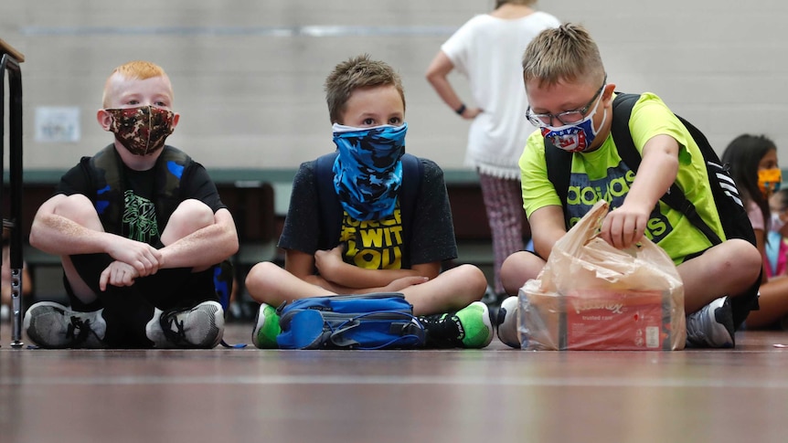 Three young boys wearing face masks and T-shirts sit on a classroom floor with other kids behind.