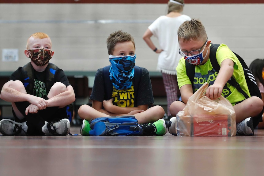 Three young boys wearing face masks and T-shirts sit on a classroom floor with other kids behind.