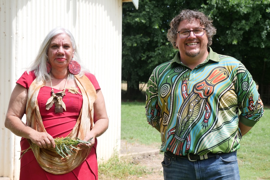 An Aboriginal woman holding leaves stands next to an Aboriginal man in front of trees and the side of a church.