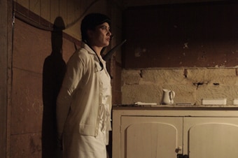 Profile or side view of woman in white uniform standing against wall in dimly lit colonial-era room, looking ahead.