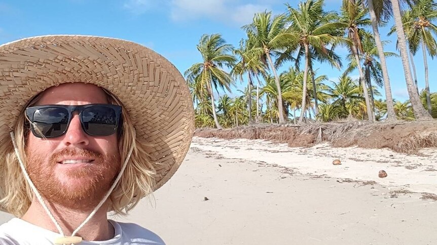 Mr Thomson wears a hat and sunglasses on a sunny beach with palm tress.