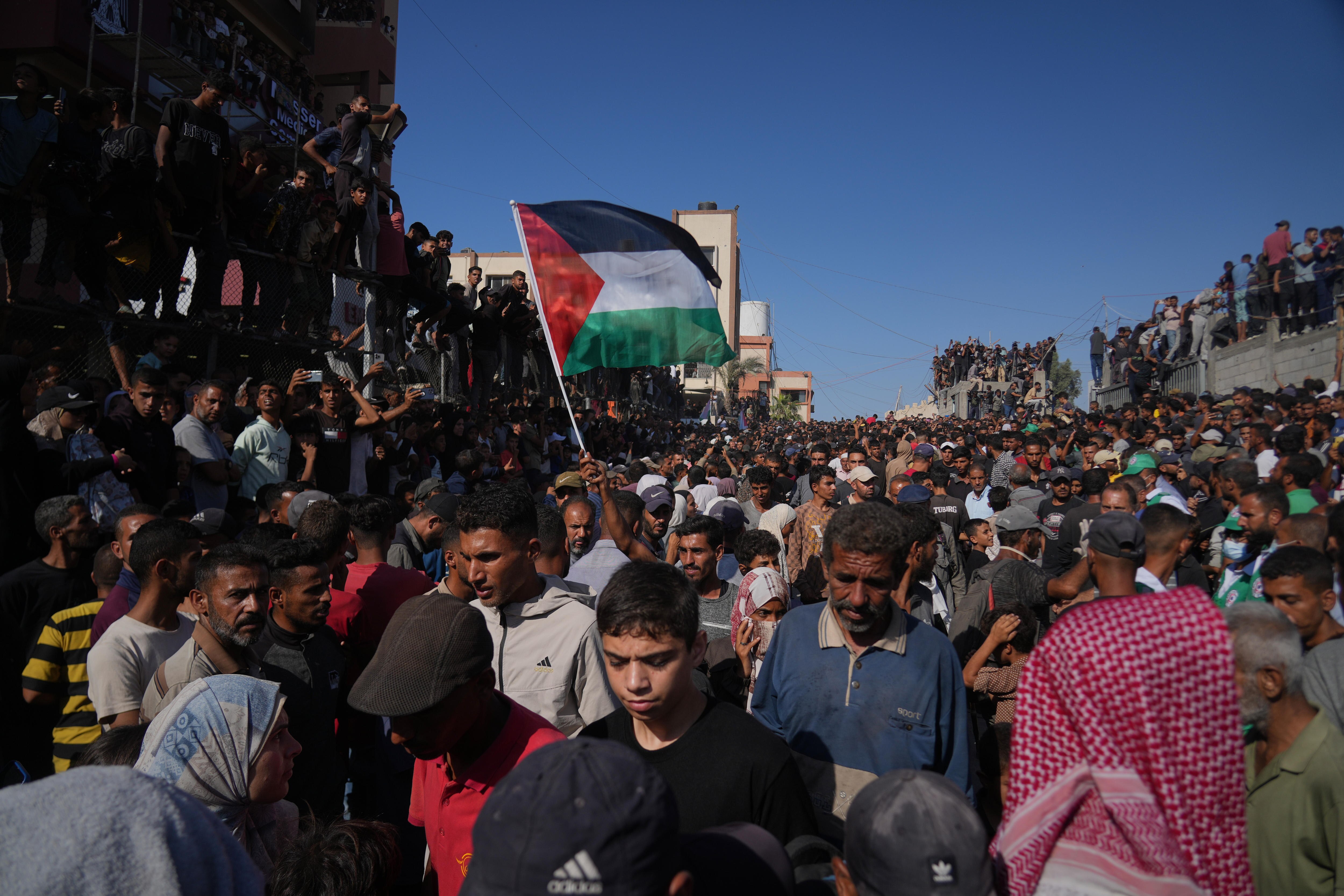 A large crowd of people standing in a street, with a single Palestinian flag raised above it.