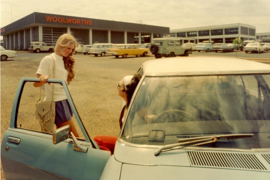 An old but colour photo of two women at a car. A shop with the word Woolworths is in the background.