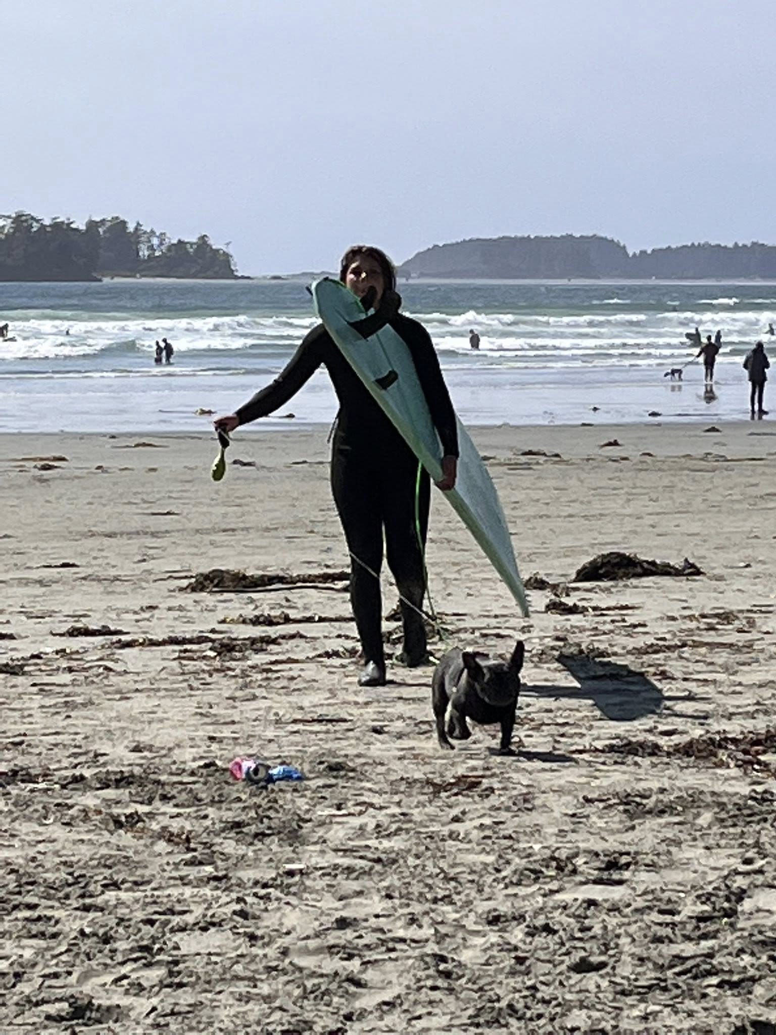 Una mujer joven con una tabla de surf bajo el brazo y un perro a su lado en la playa.