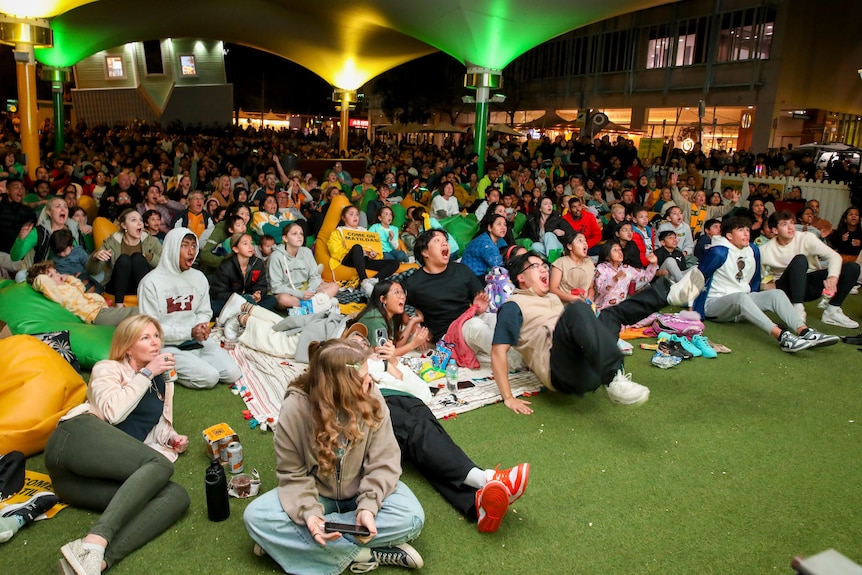 A crowd of hundreds at night shout at a big screen