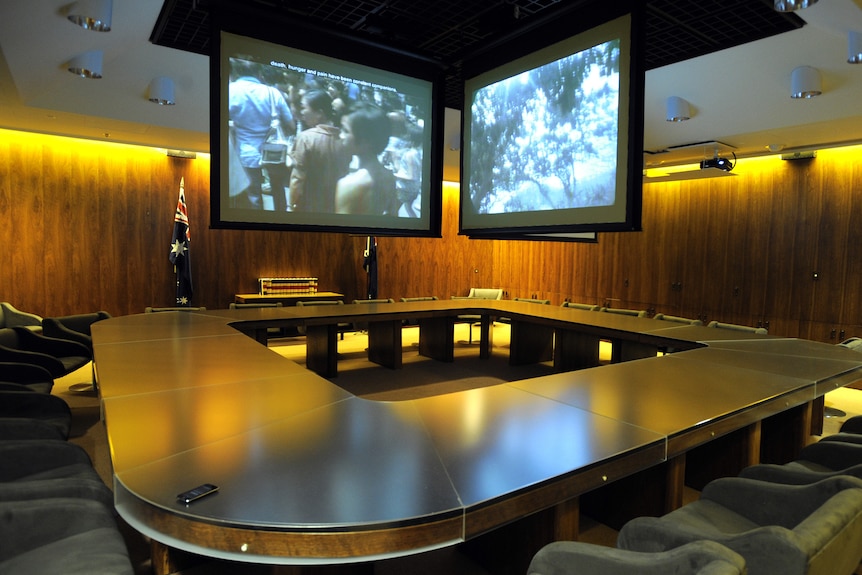A large, rounded square table sits in the middle of a posh-looking, mood-lit room below a set of video screens.