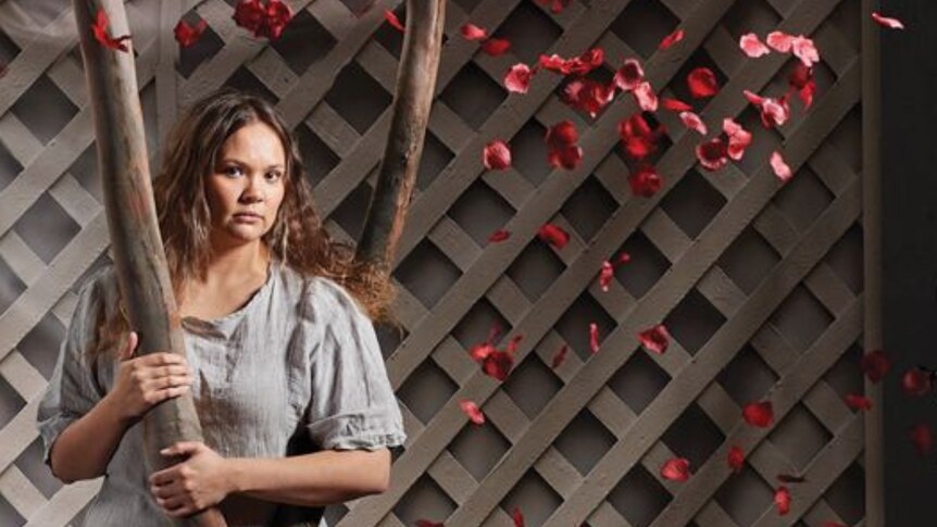 A woman stands staring at the camera, holding a tree trunk against a white lattuce fencing background with red-pink flowers