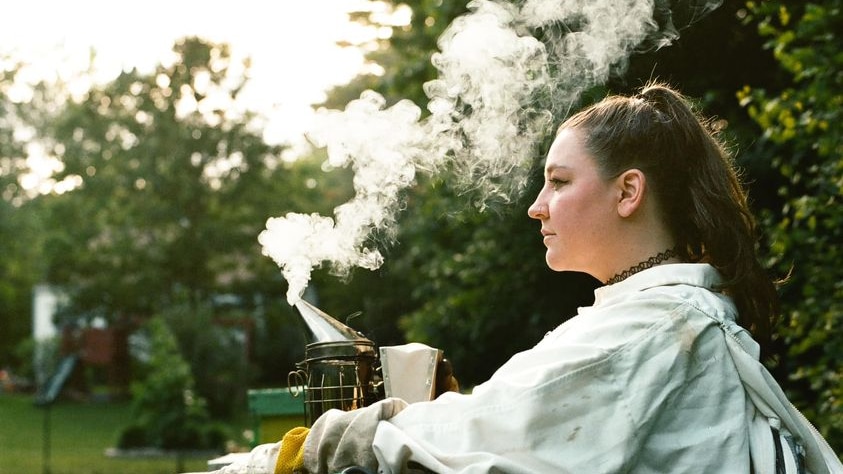 A profile photo of a woman with a brown ponytail as smoke rises from her bee smoker.