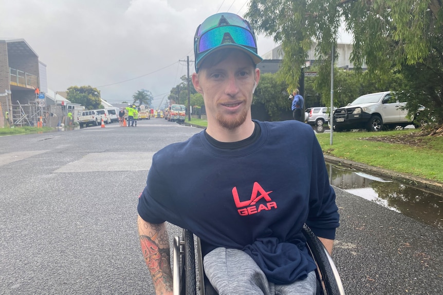 A young man in a wheelchair in a street with fire engines in the distance.