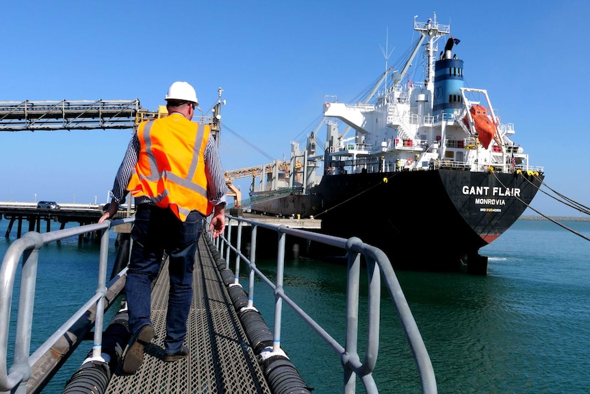 A man in a high-visibility vest and hard helmet walking up a ramp towards a cargo ship.