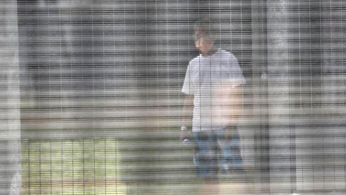 A man wearing a face mask stands behind a wire fence wearing a white shirt and jeans at Christmas Island Detention Centre.