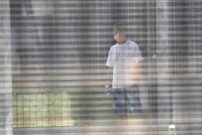 A man wearing a face mask stands behind a wire fence wearing a white shirt and jeans at Christmas Island Detention Centre.