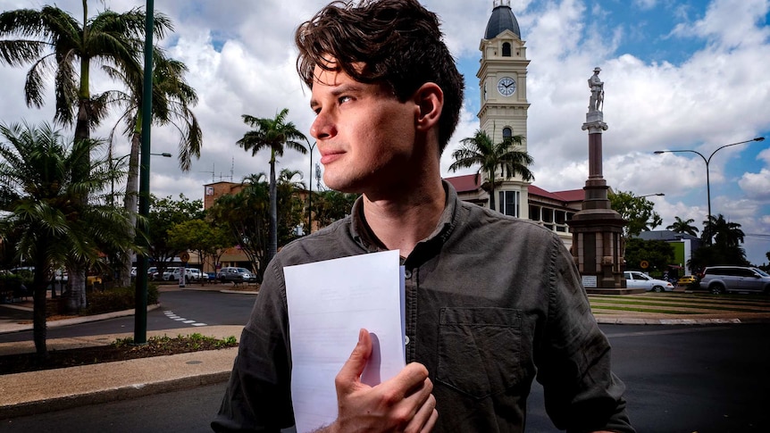 A young man in a grey shirt stands on a street in front of a clock tower, holding a folder.