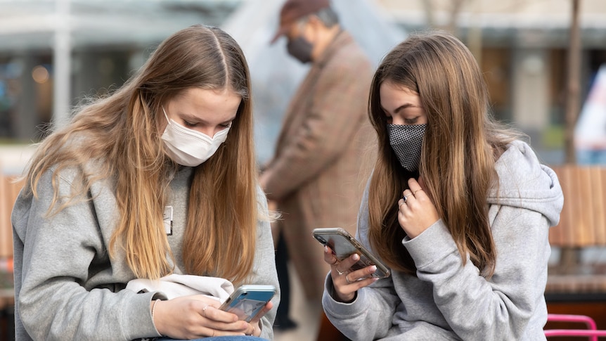 Two teenage girls on their phones sitting close together while wearing masks