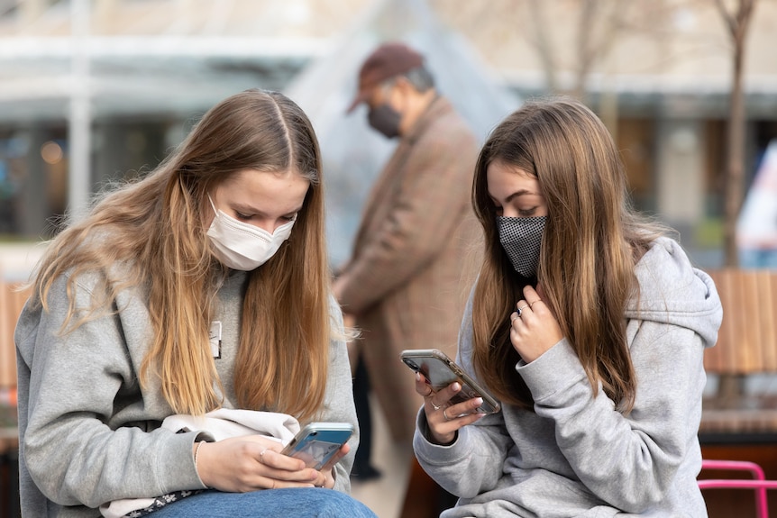 Two teenage girls on their phones sitting close together while wearing masks