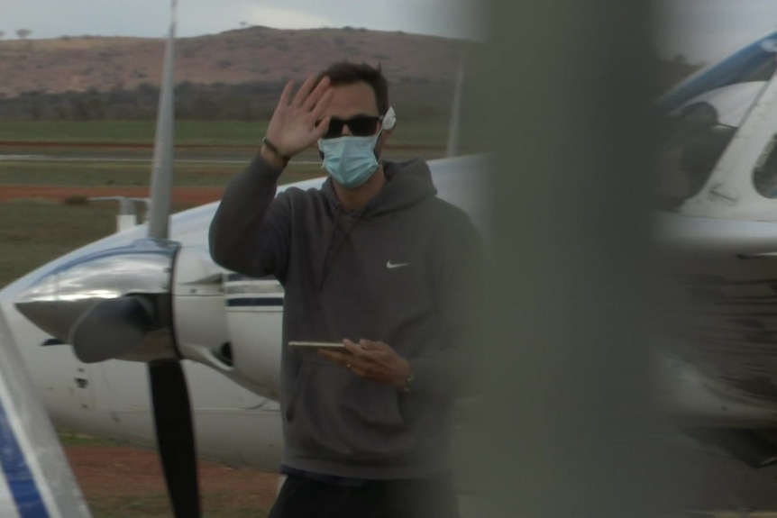 A man wearing a face mask and sunglasses next to a small plane waves at the camera 