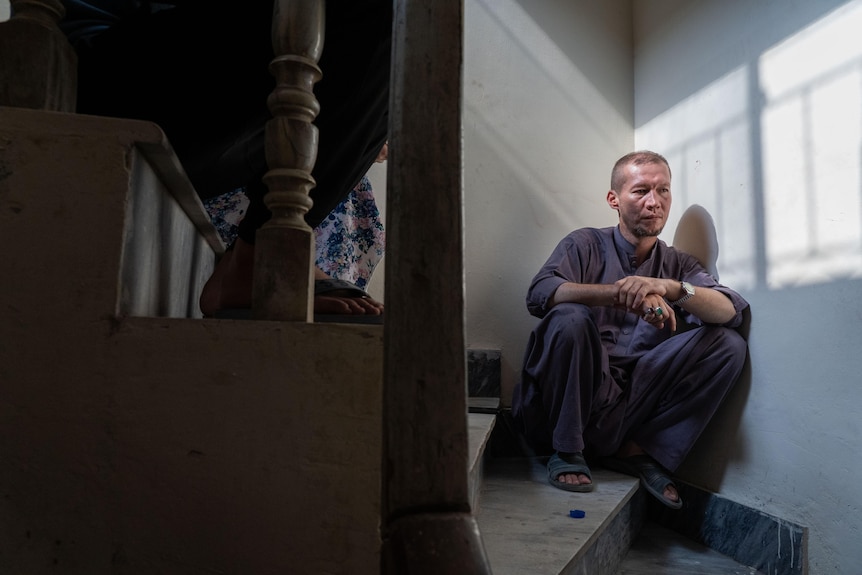 A man in blue clothes sits in a shadowy stairwell. 