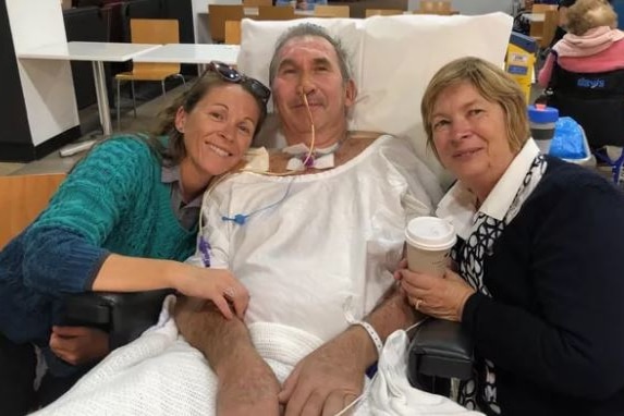 Two women cuddle a man who is being treated in hospital.