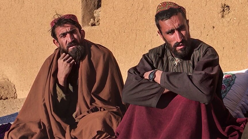 Two bearded men wearing robes and hats, sitting in front of a light brown building wall.