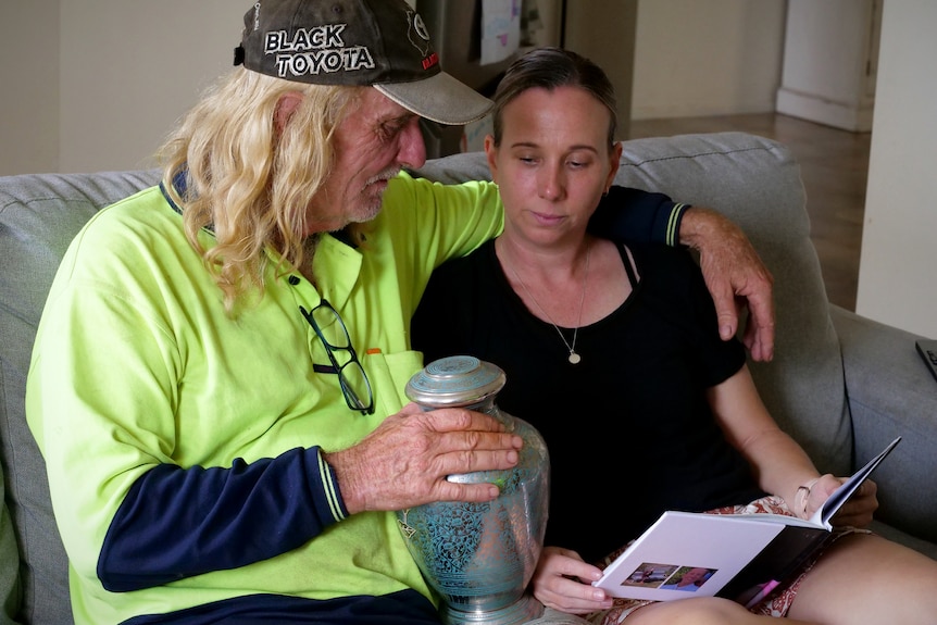 A man wearing high vis sits on a couch with his arm over a younger woman's shoulder. He is holding an urn