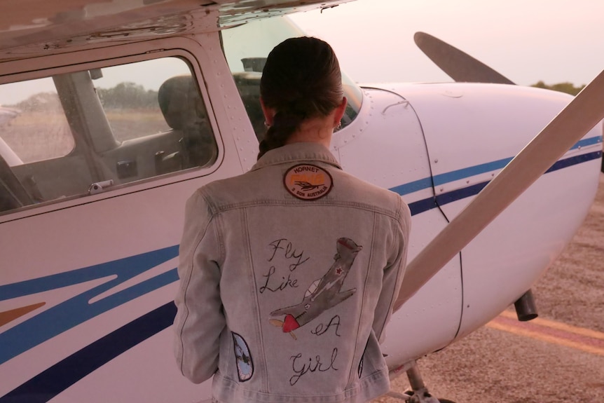 A young woman stands next to a light aircraft wearing a jacket embroidered with the phrase "fly like a girl". 