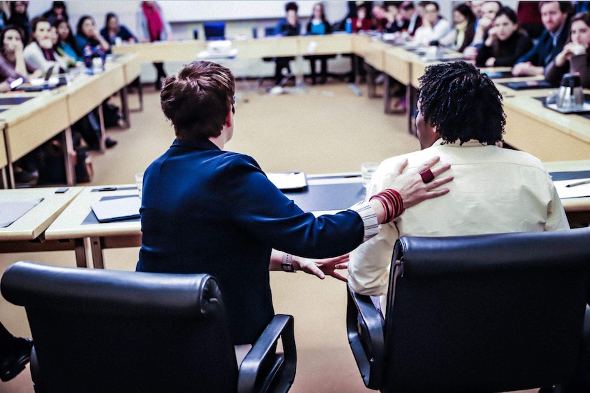 Backshot of a man and woman in a formal rectangular office full of onlookers.