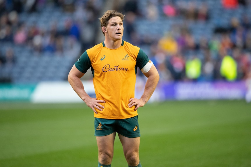 An Australian rugby player stands on the pitch with his hands on his hips.