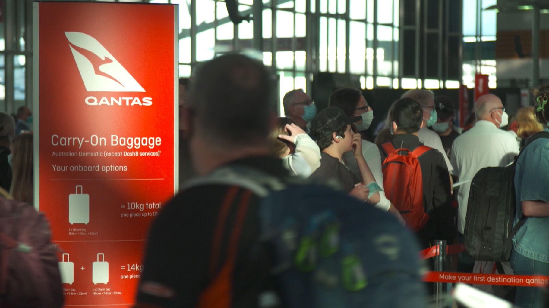 People wearing face masks queue near a Qantas sign with rules for carry-on baggage.