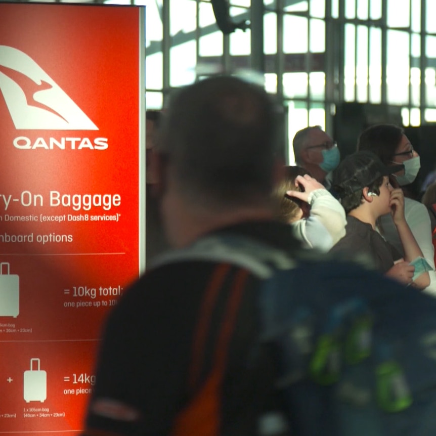 People wearing face masks queue near a Qantas sign with rules for carry-on baggage.