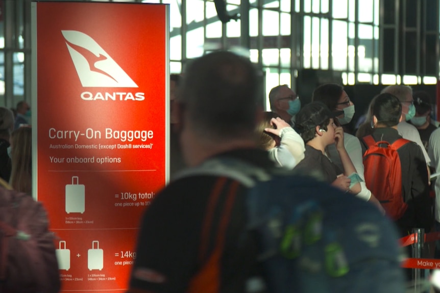 People wearing face masks queue near a Qantas sign with rules for carry-on baggage.