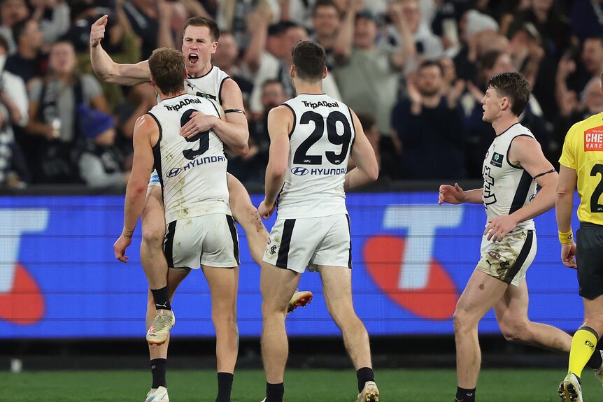 A Carlton AFL shouts with joy as his teammate lifts him up in celebration after a goal.