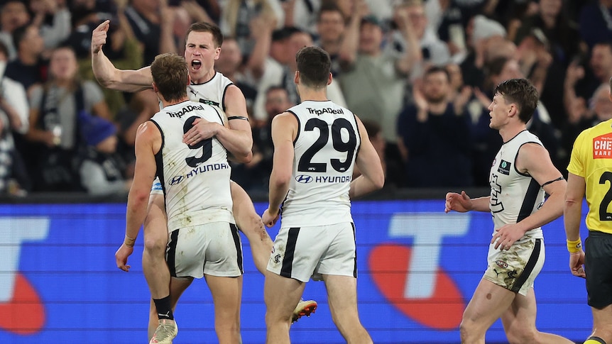 A Carlton AFL shouts with joy as his teammate lifts him up in celebration after a goal.