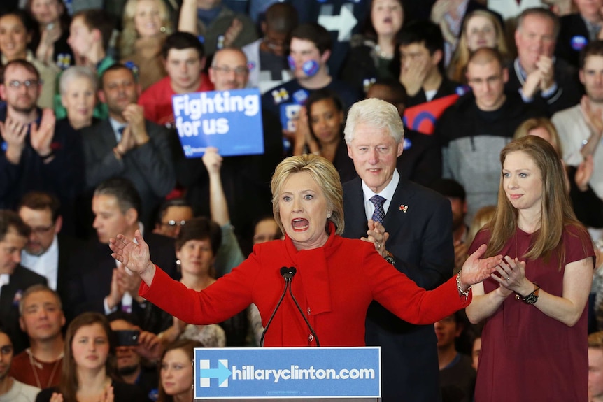 Hillary Clinton on stage, addressing supporters, as Bill Clinton and daughter Chelsea Clinton look on.