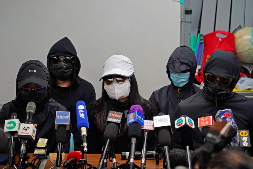 Five people wearing sunglasses with hoods over their heads sit behind rows of microphones in a dimly-lit room.