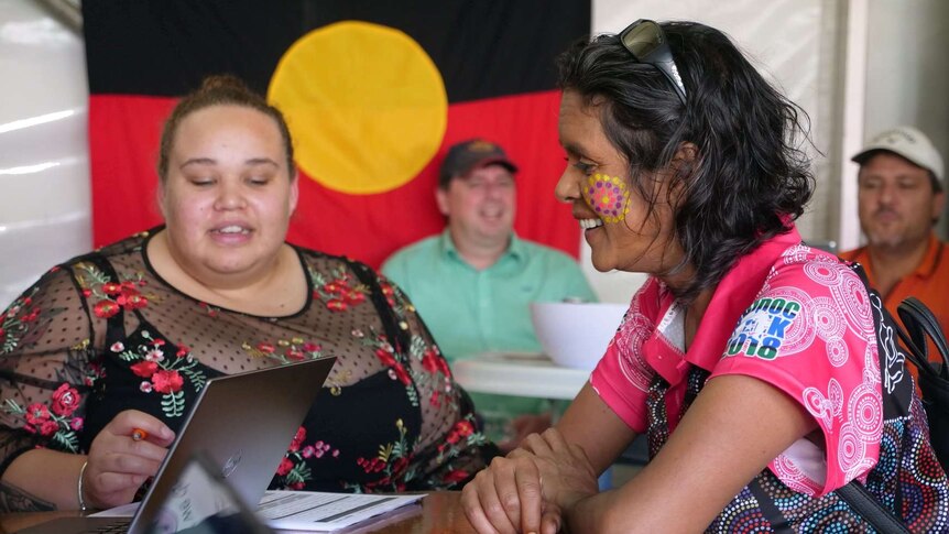 Two women sitting in front of Aboriginal flag looking at lap top