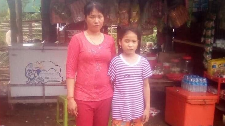A woman in a pink shirt stands next to her daughter in front of a corrugated iron hut.