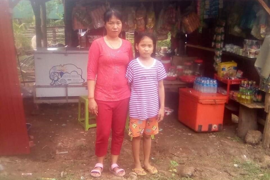 A woman in a pink shirt stands next to her daughter in front of a corrugated iron hut.