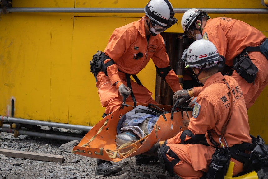 Three people in orange emergency gear and white helmets work together to carry a stretcher out of a small opening