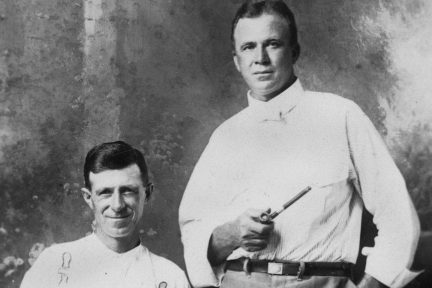 A black and white portrait of a man sitting in chair, with another man standing next to him, holding a smoking pipe in his hand.