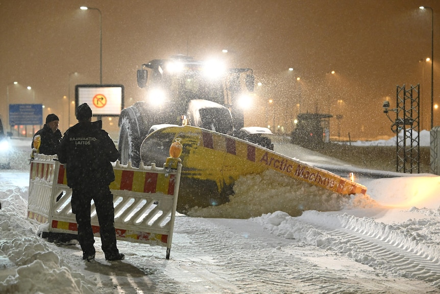 Two border guards move a temporary fence as a snow clearing truck clears a road