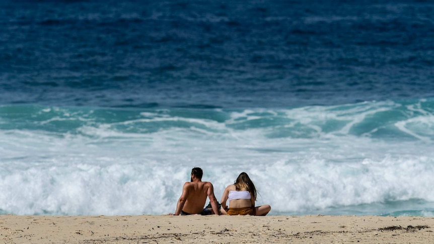 A couple sunbake while sitting on the beach, against a backdrop of blue ocean.