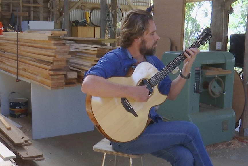 Guitarist sitting and playing  in the middle of a wood-worker's shed.